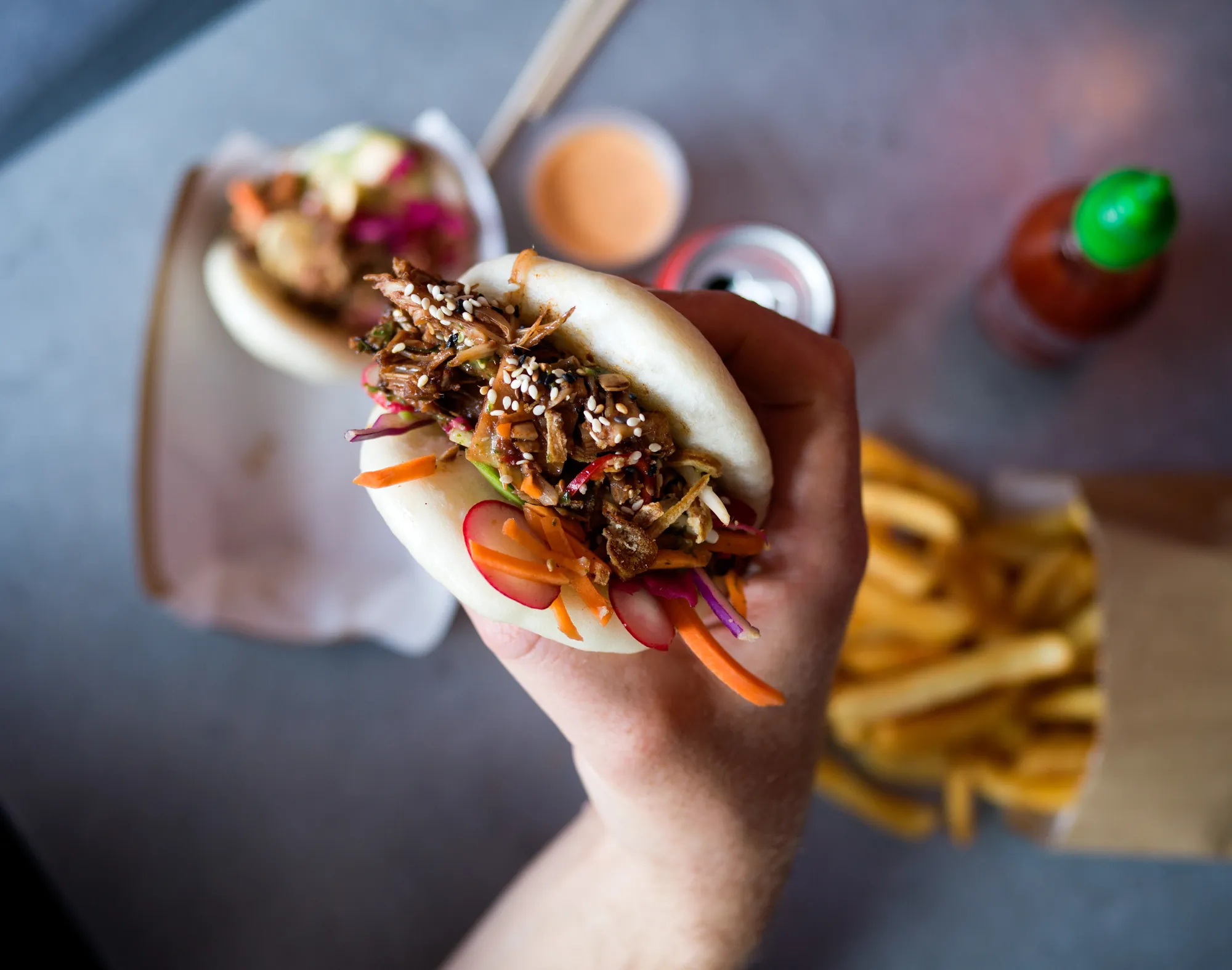 Close-up of a hand holding a bao bun with pulled filling, slaw and sesame seeds
