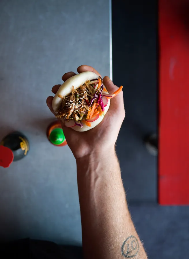 Hand holding a bao bun with pulled filling, slaw and sesame seeds over a grey table