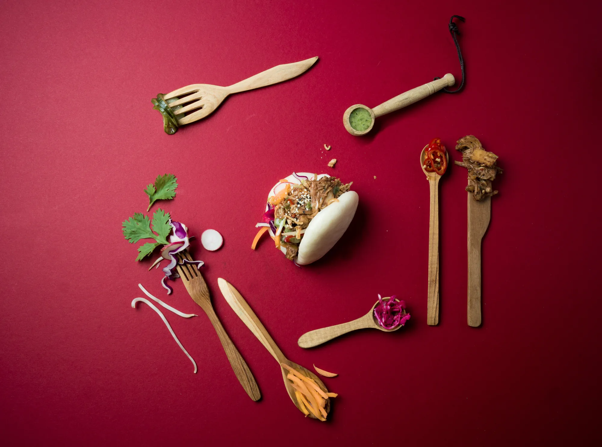 Flat lay of a bao bun surrounded by wooden spoons holding its fresh ingredients on a red background