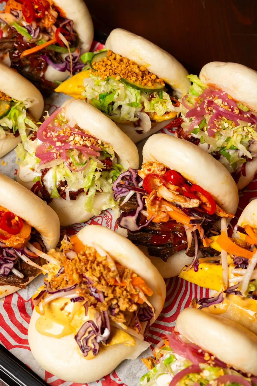 Platter of steamed bao buns on Bao Bun branded paper, filled with pulled meat, crispy chicken, slaw, red cabbage, pickled red onion, red chillies, crushed peanuts and crispy shallots