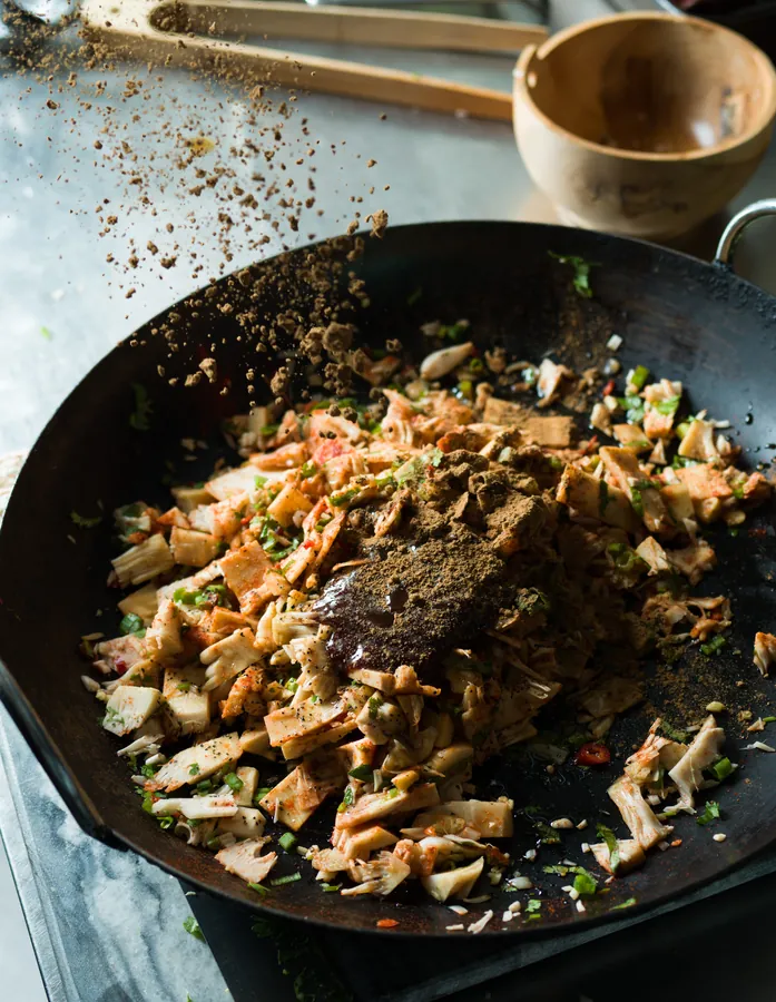 Spices being sprinkled over a shredded filling in a black wok