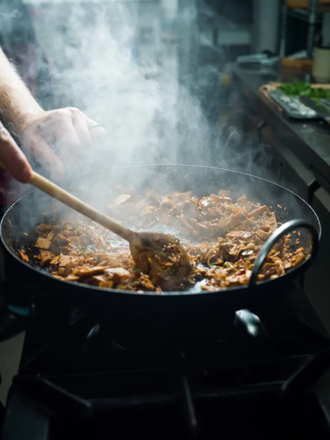 Chef stirring a shredded filling in a steaming wok on a gas hob